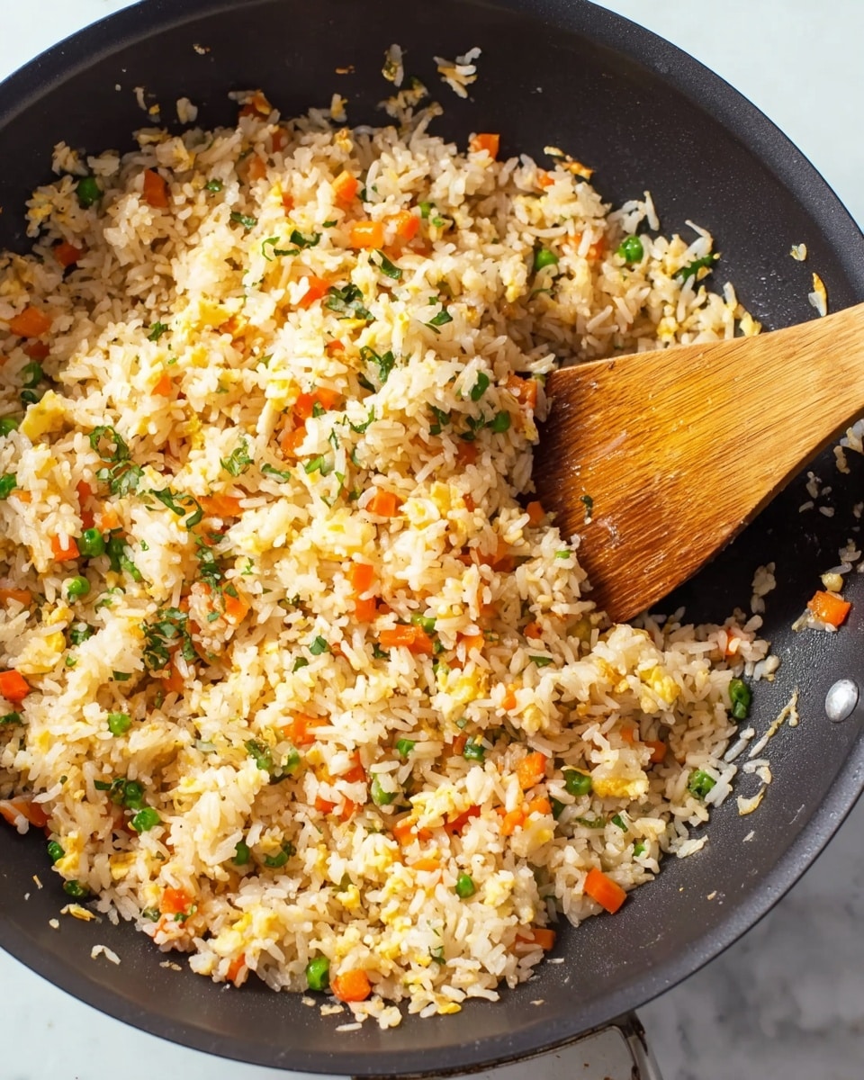 A close-up view of a black frying pan filled with cooked fried rice mixed evenly with small pieces of orange carrot and green peas. The rice looks fluffy and has a light golden color, with bits of scrambled egg scattered throughout. There is a wooden spatula resting inside the pan, slightly lifting the rice from one side. The pan is placed on a white marbled surface that adds a clean and bright background to the dish. photo taken with an iphone --ar 4:5 --v 7