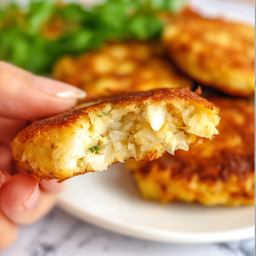 A close-up shows a golden-brown fried patty held by a woman's hand, revealing its soft, fluffy inside filled with small pieces of light-colored onions and a slightly spongy texture. In the blurred background, more patties rest on a white plate, with a green leafy garnish behind them, all set on a white marbled surface. The lighting is bright, highlighting the crispy edges and the tender inside of the patty photo taken with an iphone --ar 4:5 --v 7