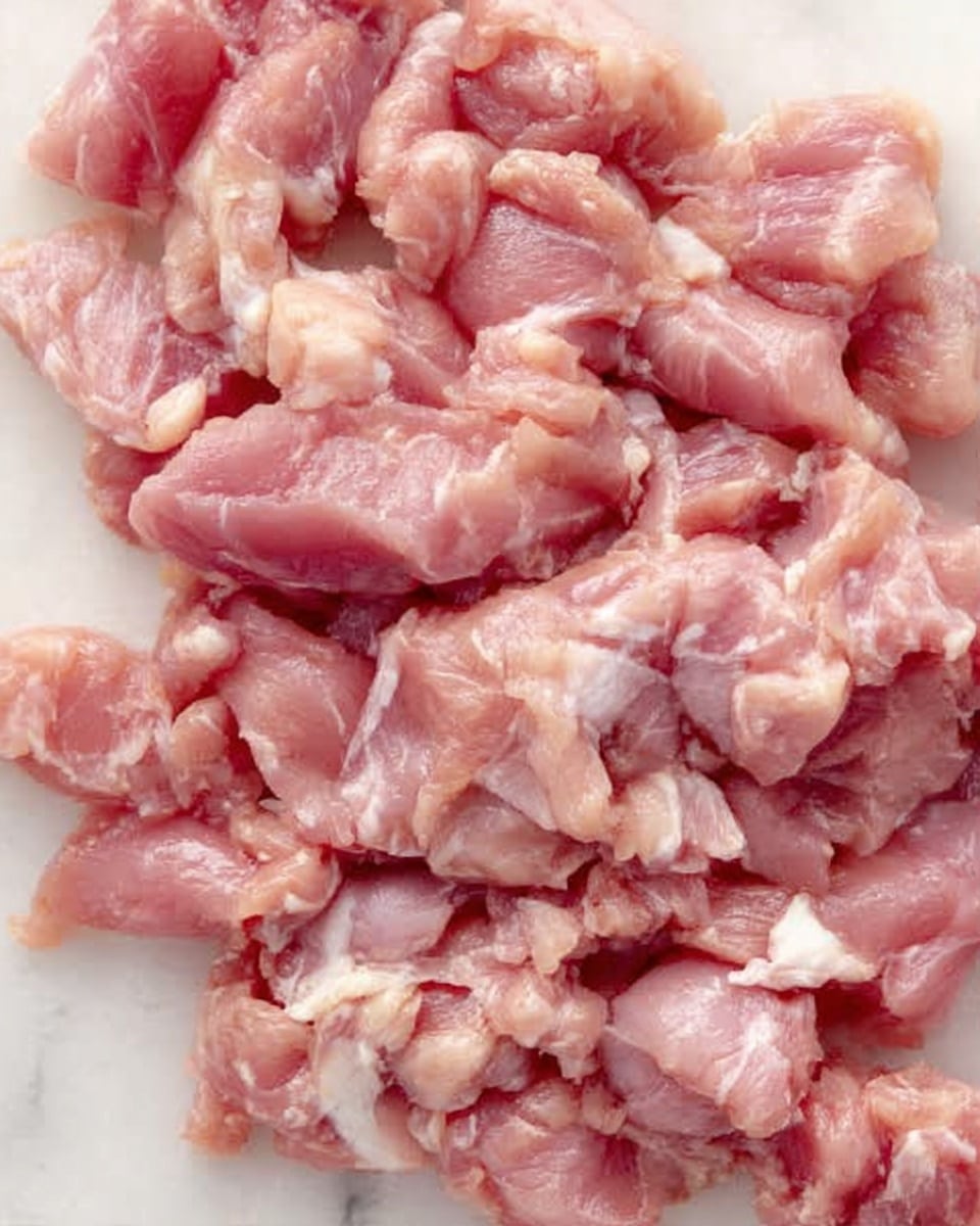 The image shows a close-up of many pieces of raw pink chicken meat lying scattered on a smooth white marbled surface. The chicken pieces vary in size and have some white fat parts mixed in. The overall texture appears soft and slightly shiny from the freshness of the meat. There are no other items or colors in the image, and no visible hands or utensils. photo taken with an iphone --ar 4:5 --v 7