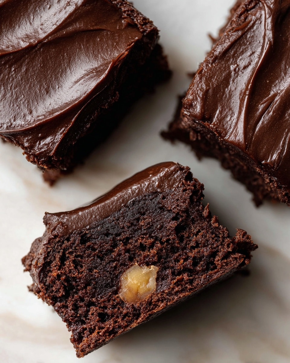 The image shows three close-up pieces of dark chocolate cake with a glossy, smooth layer of dark chocolate frosting on top. One piece is cut to show the inside, which has a rich, moist, dark brown texture with a small visible chunk of light brown or tan-colored nut or fruit embedded in it. The cakes are placed on a white marbled surface, making the dark chocolate color stand out clearly. The frosting has subtle swirls and a shiny finish, with a soft and dense crumb texture visible inside the cut piece. photo taken with an iphone --ar 4:5 --v 7