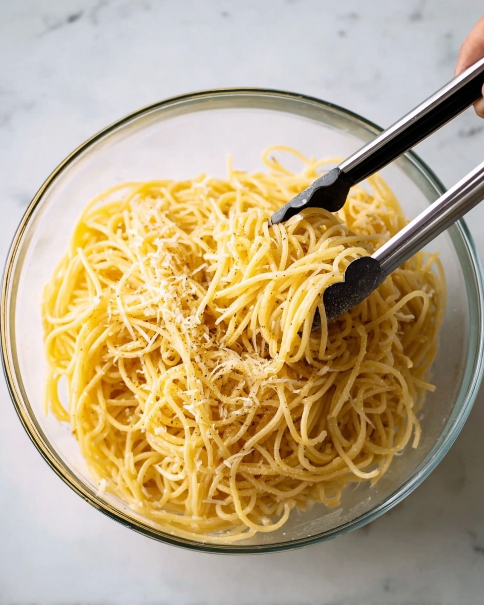 A clear glass bowl filled with cooked spaghetti pasta that is light yellow in color, tossed with a sprinkling of black pepper and grated cheese, giving it a slightly textured surface. The spaghetti strands are loosely piled with some strands lifted by black-handled metal tongs held by a woman's hand, positioned above the bowl. The bowl sits on a white marbled surface, creating a clean, simple background. Photo taken with an iphone --ar 4:5 --v 7