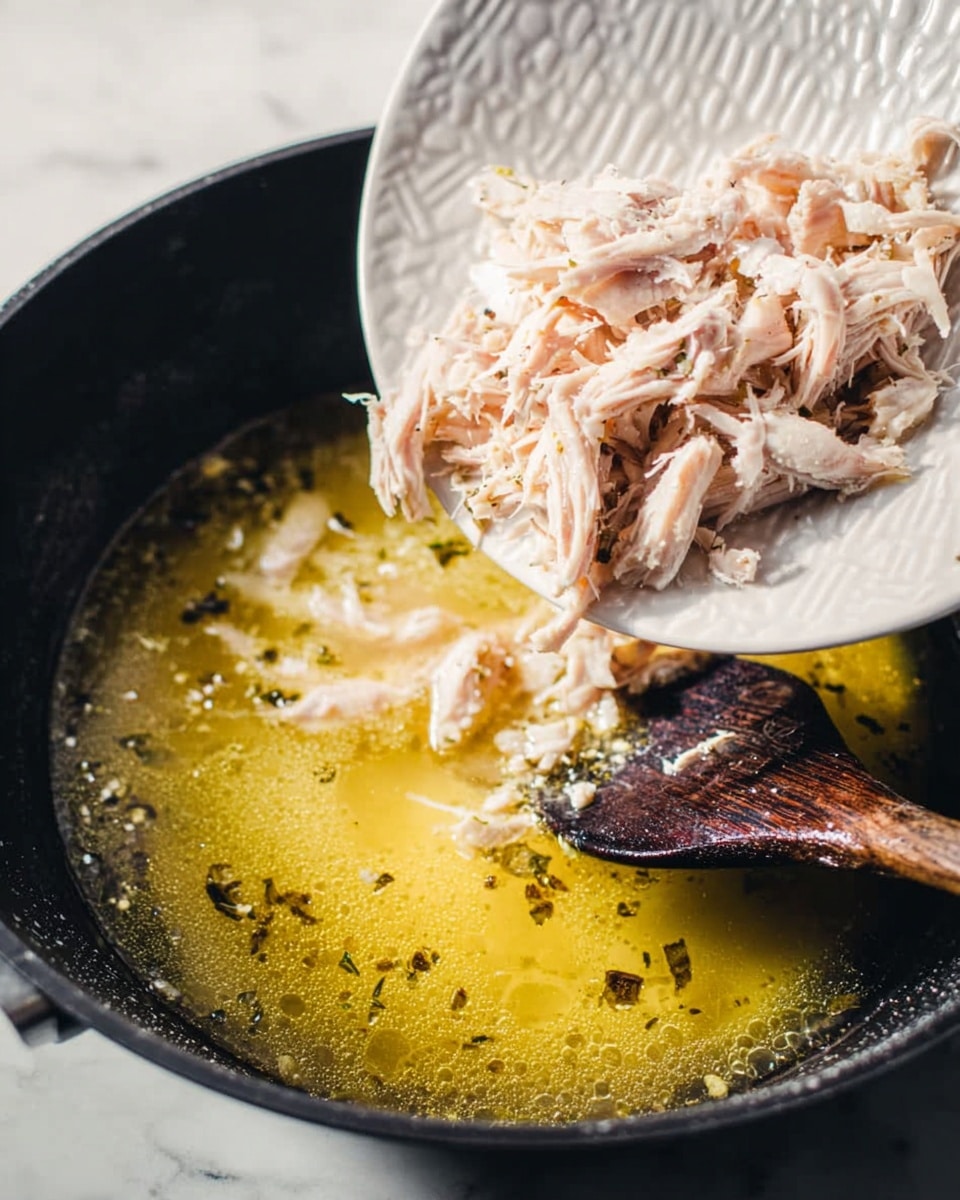 The image shows shredded light pink chicken being dropped from a white plate with a textured pattern into a black pot filled with yellowish broth that has small bubbles and herbs floating on the surface. A dark wooden spoon is partly visible under the shredded chicken on the plate. The pot is on a white marbled surface. The photo taken with an iphone --ar 4:5 --v 7