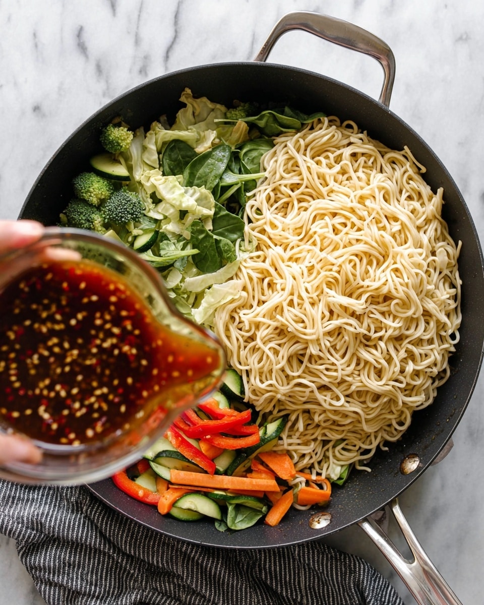 A large black pan is shown from above on a white marbled surface with a grey and black striped cloth beneath; inside the pan, on the right side, there is a big pile of light yellow cooked noodles with a smooth texture, while on the left side, there is a colorful mix of fresh vegetables including bright green broccoli florets, dark green spinach leaves, light green cabbage pieces, green zucchini slices, red bell pepper strips, and orange carrot sticks. In the lower left corner, a woman's hand holds a small glass measuring cup pouring a reddish-brown sauce with visible red chili flakes and seeds onto the vegetables. The pan has a silver metal handle, and the photo is bright with soft shadows. Photo taken with an iphone --ar 4:5 --v 7