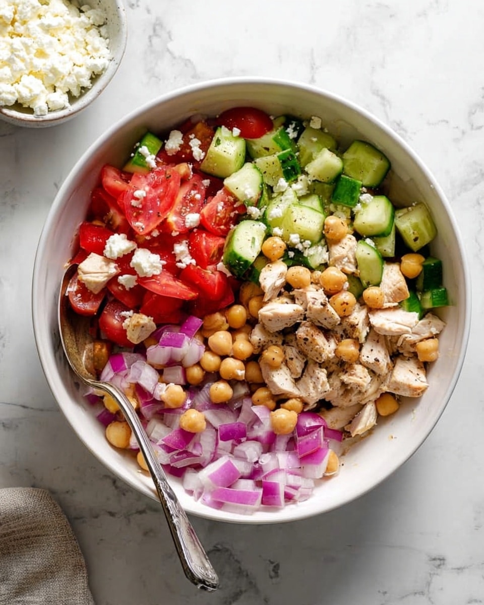 The image shows a white bowl filled with a colorful salad on a white marbled surface. Inside the bowl, the salad has several layers: at the base are small pieces of red onion scattered around, followed by a mix of round, light brown chickpeas evenly spread over the salad. On top, there are chopped green cucumbers with their rough skin and chunks of bright red tomatoes. Pieces of light beige cooked chicken are placed on top, for a final layer, mixed evenly among the vegetables and chickpeas. A silver spoon rests inside the bowl, leaning on the edge. To the side, a small bowl with white crumbled cheese is partly visible. Photo taken with an iphone --ar 4:5 --v 7
