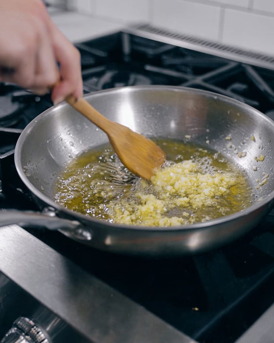A silver metal pan sits on a black stove burner, with light yellow garlic pieces being stirred in hot oil inside the pan. A woman's hand is holding a wooden spoon, moving it to mix the garlic and oil. The garlic is finely chopped, floating and sizzling in the clear, golden oil, creating small bubbles around it. The background shows part of the stove with control knobs and a white tiled wall. photo taken with an iphone --ar 4:5 --v 7