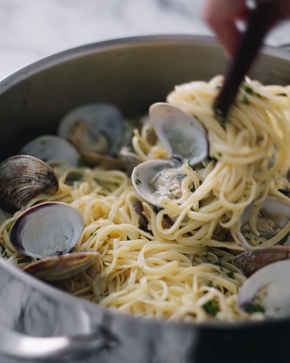 The image shows a close-up of a large silver pot filled with cooked long noodles mixed with clams, some still inside their shells, and herbs. The noodles are light yellow and soft, tangled together with small green herbs sprinkled throughout. A woman's hand with a dark utensil is gently lifting some noodles and a clam from the pot. The background has a white marbled texture. photo taken with an iphone --ar 4:5 --v 7
