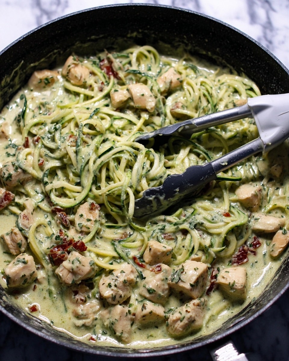 The image shows a black pan filled with creamy green sauce pasta made with spiralized zucchini noodles that are light green with a smooth wet look. The sauce coats the noodles and cubed light beige chicken pieces mixed evenly throughout. Small bits of dark green herbs are spread all over the dish along with some dried red tomato pieces adding color contrast. A pair of gray and black tongs and a white spatula with a dark handle rest inside the pan, partly covered by the sauce. The background is a white marbled texture. photo taken with an iphone --ar 4:5 --v 7