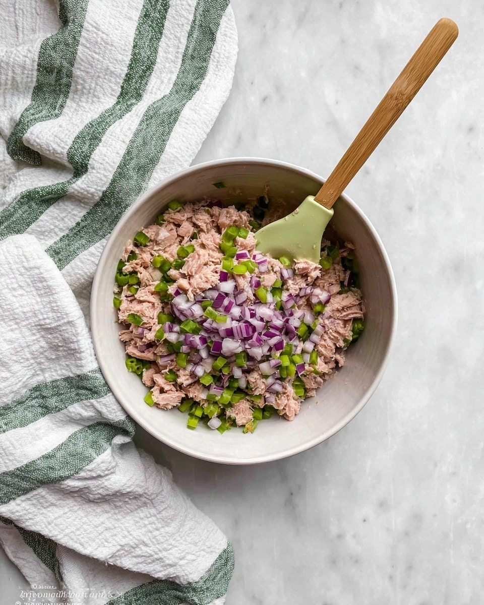 In a white bowl placed on a white marbled surface, there is a mix of three main layers: the bottom layer consists of chunks of light brown tuna fish; lightly scattered on top are small pieces of green celery; and finely chopped pieces of purple and white red onion are spread evenly over the celery. A wooden spoon with a light green silicone head is resting inside the bowl, partly covered by the tuna mix. A white cloth with green stripes lies crumpled near the bottom left of the bowl. photo taken with an iphone --ar 4:5 --v 7