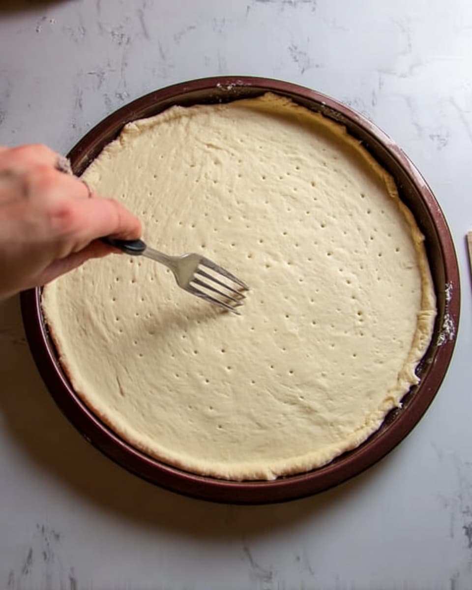 A round pizza dough spread out evenly inside a dark brown pizza pan, resting on a white marbled surface. The dough is pale cream in color with a smooth texture and small holes being made on the surface by a silver fork held by a woman's hand coming from the top left. The dough edges are slightly thicker and uneven. photo taken with an iphone --ar 4:5 --v 7