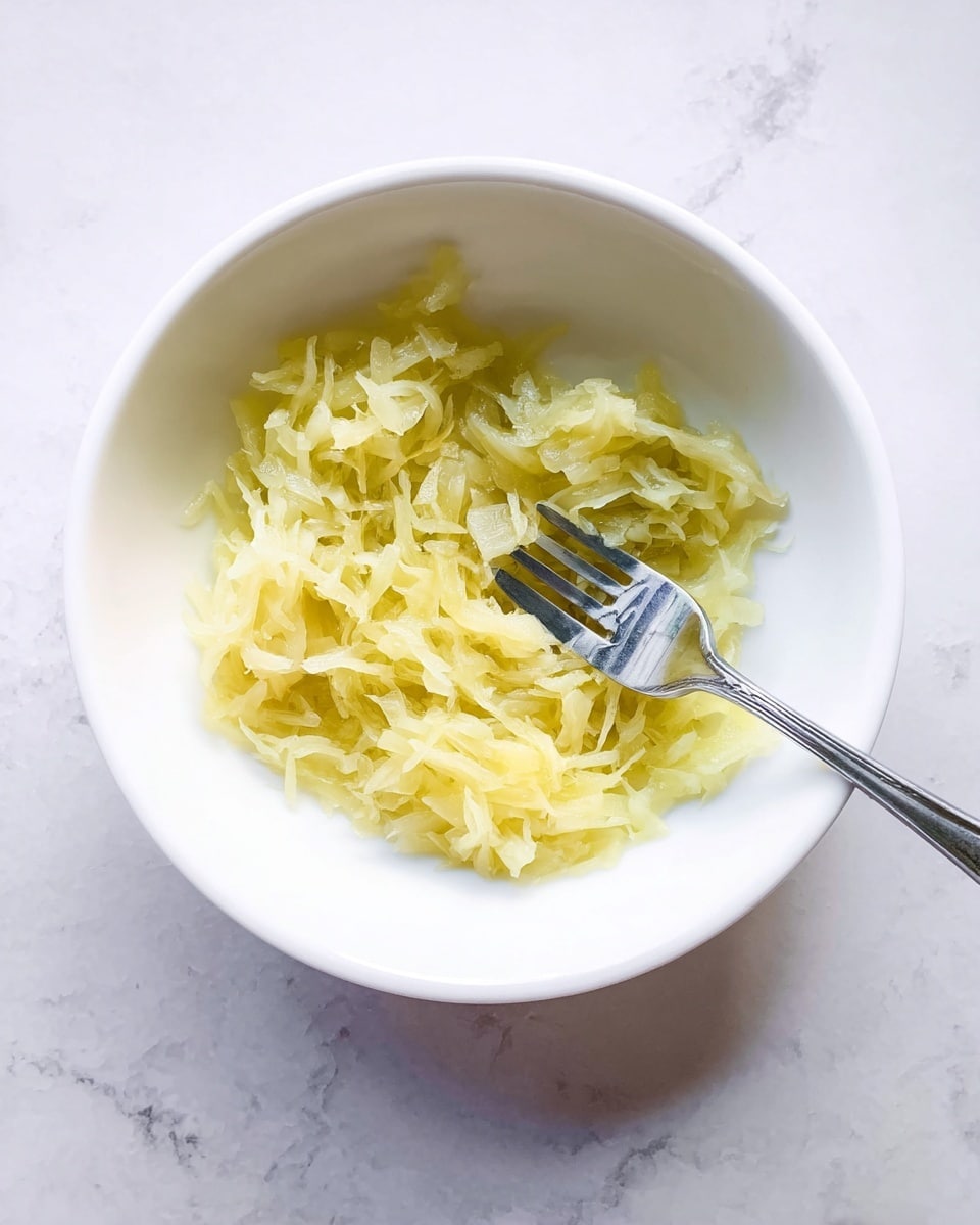 A white bowl filled with one layer of pale yellow shredded fruit or vegetable, soft and moist in texture. Inside the bowl, a shiny silver fork rests on the shredded pieces, with some of the shreds slightly pushed aside, showing the smooth white surface of the bowl underneath. The bowl sits on a white marbled surface with faint gray veins. photo taken with an iphone --ar 4:5 --v 7