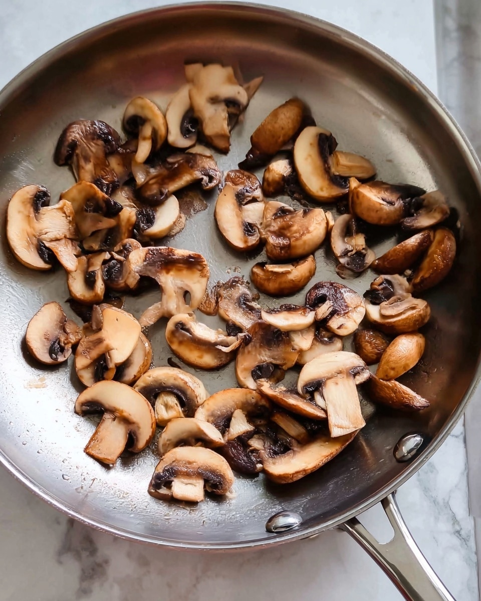 The image shows a silver frying pan filled with sliced brown mushrooms that are lightly cooked and spread out loosely across the pan. The mushrooms have a soft, slightly shiny texture with some darker brown spots indicating cooking. The background surface is a white marbled texture, and the pan's metal handle extends out towards the bottom left of the image. Photo taken with an iphone --ar 4:5 --v 7