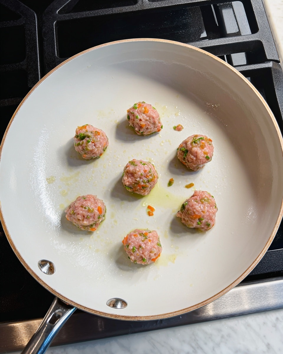 A white pan with a slightly browned rim holds eight small, round pink meat mixtures spaced evenly inside. Each small ball has visible pieces of green and orange bits mixed in. Light drops of oil are scattered on the pan surface, which has a smooth and clean white interior. The pan handle is metallic silver, and the pan sits on a stove with a dark burner area and a white marbled countertop visible below. Photo taken with an iphone --ar 4:5 --v 7