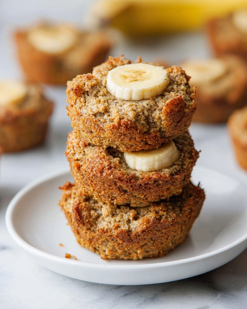 A stack of three thick, round banana muffins with a rough, crumbly texture is placed on a white plate. Each muffin is golden brown with uneven, rustic edges, and the top muffin has a small, round slice of banana embedded in its center. In the background, more muffins with the same features are scattered on a white marbled surface. The image is softly lit with a shallow depth of field, focusing on the stack of muffins. photo taken with an iphone --ar 4:5 --v 7