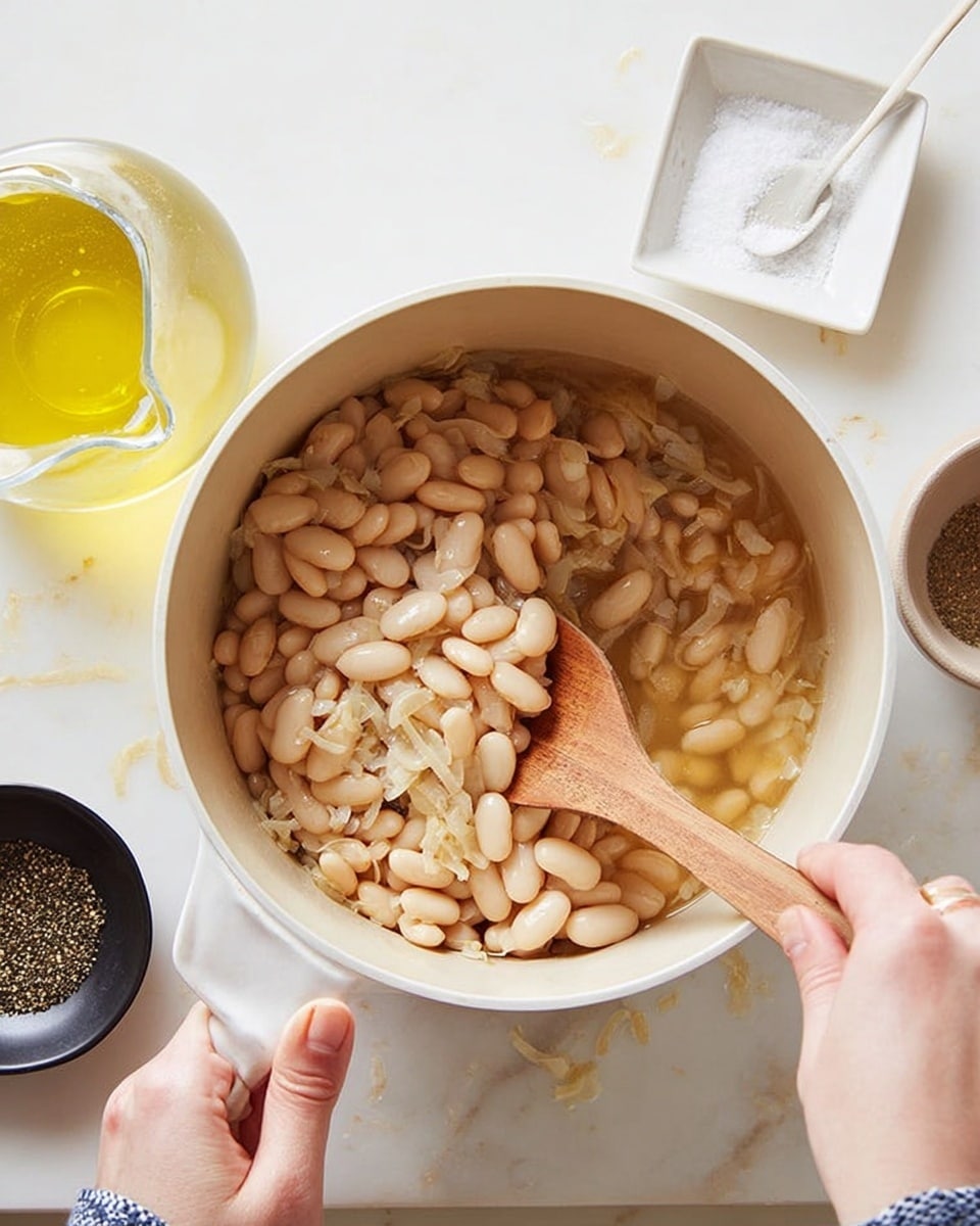 A close-up view of a white pot filled with pale beige beans, scattered with small pieces of light translucent onion and specks of black seasoning. A woman's hand holds a wooden spoon stirring the beans inside the pot, while the other woman's hand grips the pot handle. Nearby on a white marbled surface are a glass pitcher with a yellow liquid, a small white square bowl with white granulated salt and a white spoon, and a black bowl with cracked black pepper. The scene is bright and clean with soft natural lighting. photo taken with an iphone --ar 4:5 --v 7