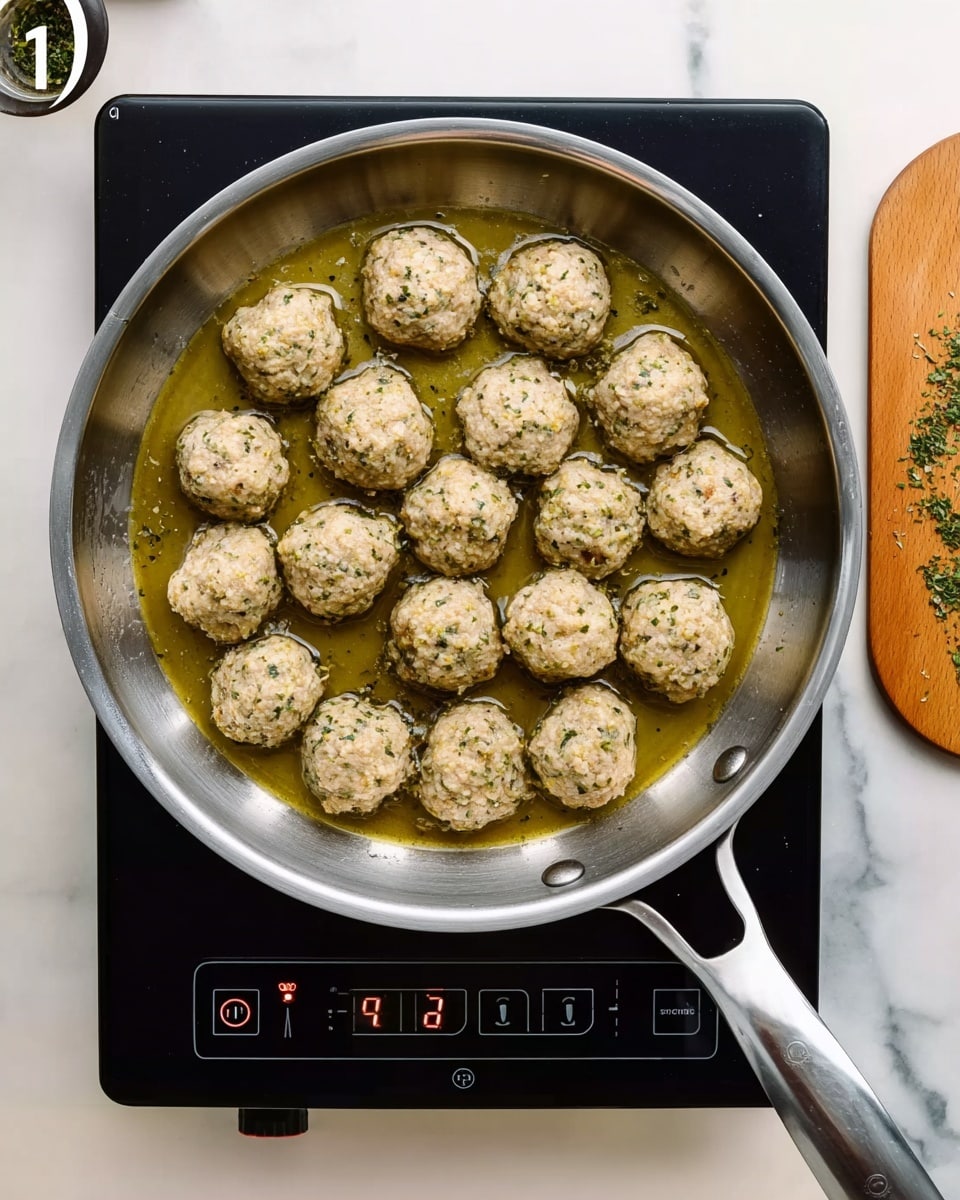 A silver frying pan sits on a black electric stovetop with 15 small, round meatballs inside, arranged in loose rows. The meatballs have a light beige coating with small green specks, resting in a shallow pool of golden oil that covers the pan's bottom. The stovetop displays a digital screen with buttons and numbers visible, and a wooden surface is seen to the right side with small green herb pieces scattered. The scene is set against a white marbled texture. photo taken with an iphone --ar 4:5 --v 7