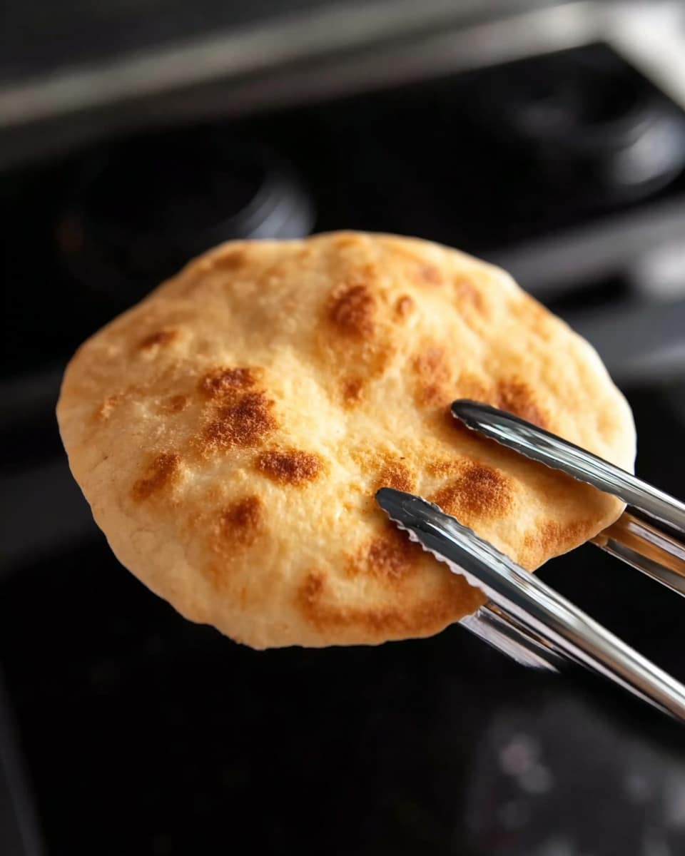 A single piece of golden brown puffed flatbread is held above a black stove by silver metal tongs, showing a smooth and slightly shiny surface with a soft, airy texture. The background has dark stove parts, and the flatbread looks freshly cooked with light browned spots on its surface, slightly puffed out with a round shape and a few gentle curves along the edges. Photo taken with an iphone --ar 4:5 --v 7