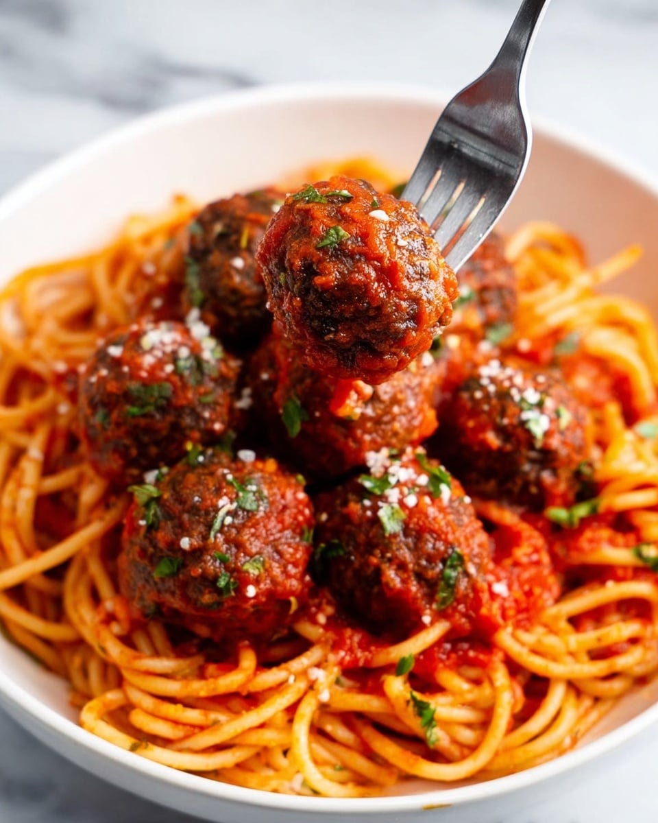 A white bowl filled with a bed of twisted, light orange spaghetti noodles at the bottom, covered with a bright red tomato sauce that looks thick and chunky. On top, there are about seven dark brown meatballs coated evenly with the same tomato sauce. Green herb bits are sprinkled on the meatballs and sauce to add color, and some small white cheese flakes add extra texture. A silver fork is holding one meatball in the air, showing its round and textured surface. The bowl is placed on a white marbled surface. photo taken with an iphone --ar 4:5 --v 7
