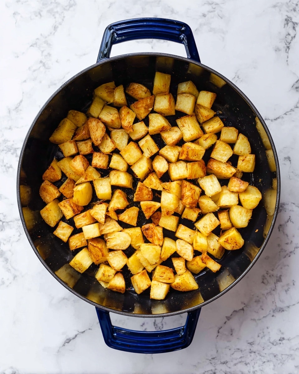 This image shows a black pan with two navy blue handles placed on a white marbled surface. Inside the pan, there are small golden-brown potato cubes with the skin still on, spread evenly across the bottom. The potatoes have a slightly crispy texture with color shades ranging from light yellow to golden brown, showing some pieces are more cooked on their edges. photo taken with an iphone --ar 4:5 --v 7