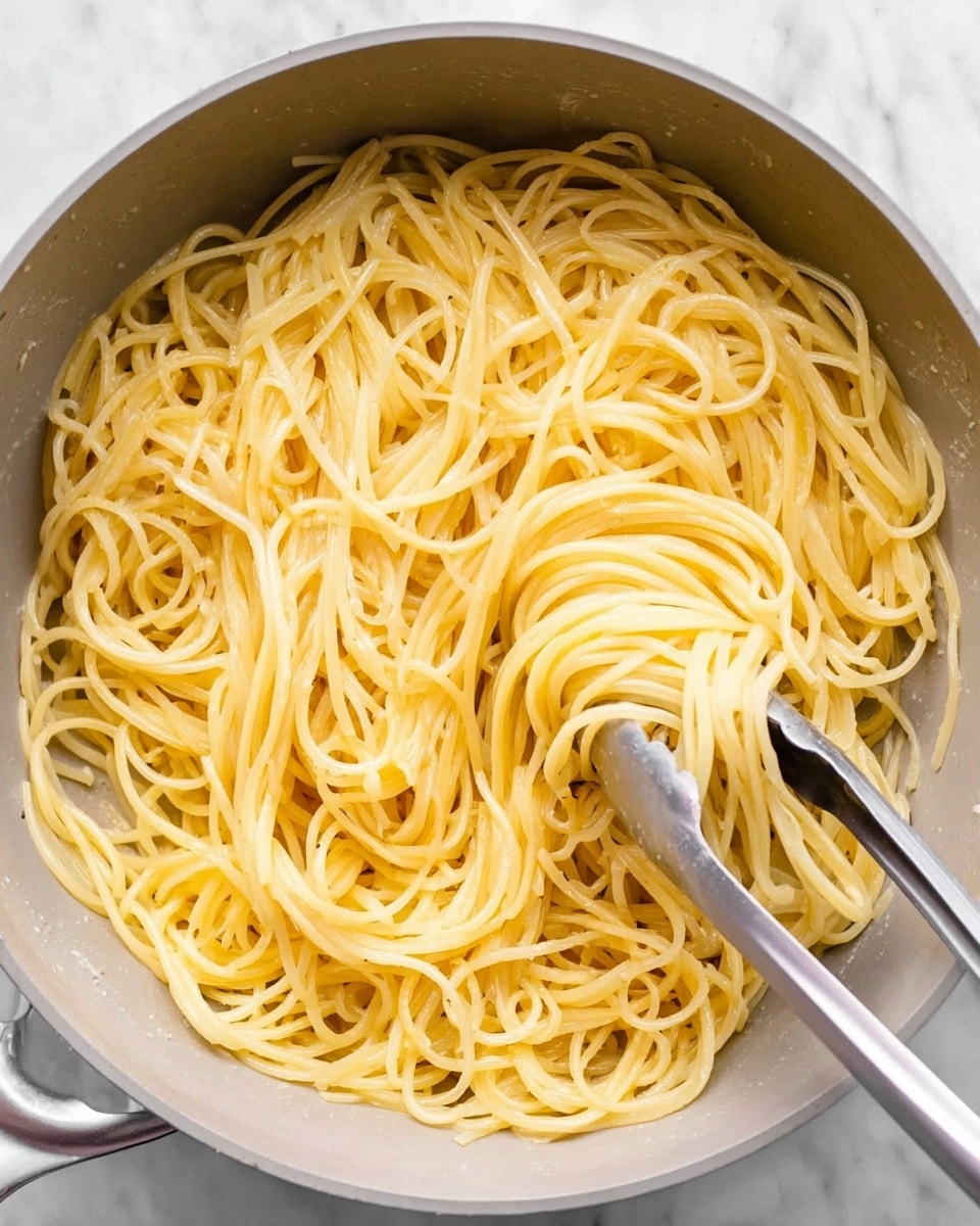 This image shows a close-up view of cooked spaghetti pasta inside a large pot. The spaghetti strands are pale yellow, soft, and slightly shiny, spread loosely all around with some strands curling and overlapping each other. A pair of silver metal tongs holds a bundle of the spaghetti in the center right of the pot, lifting it slightly above the others. The pot is light gray inside with a smooth texture. The background surface is a white marbled texture. photo taken with an iphone --ar 4:5 --v 7