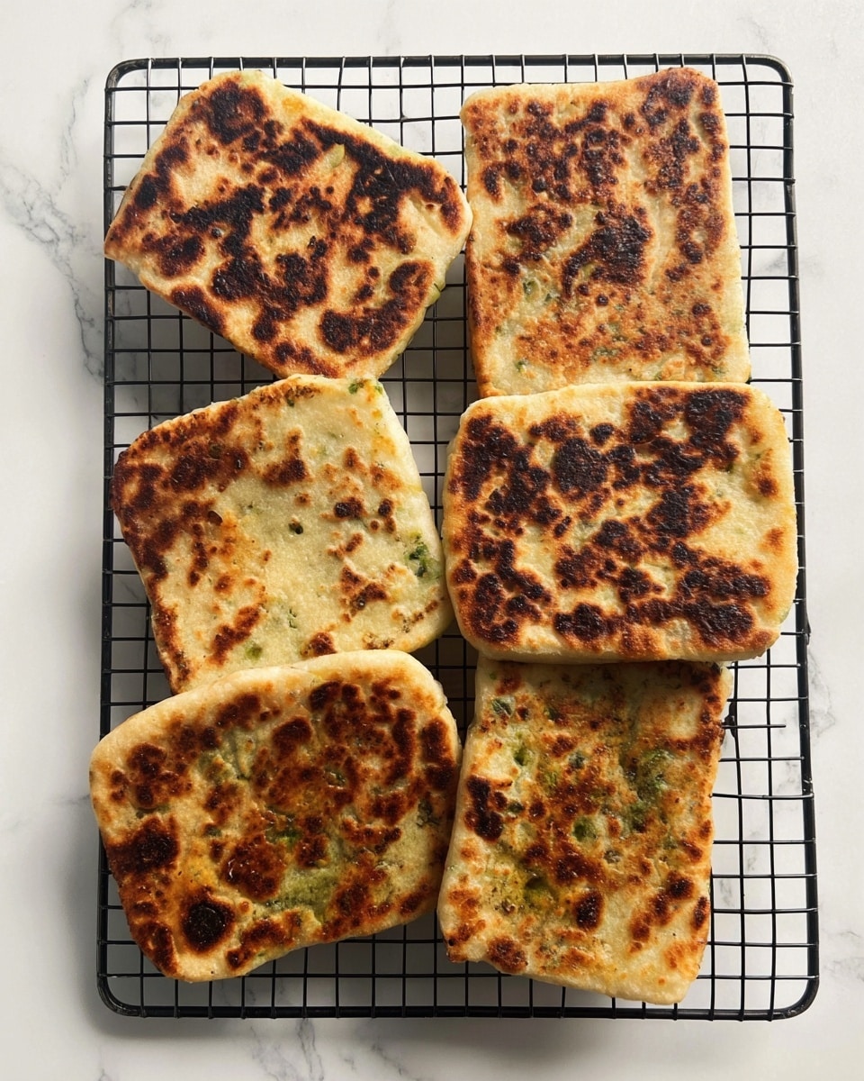 The image shows six square-shaped flatbreads with golden to dark brown charred spots, placed on a black wire cooling rack. Each flatbread has a slightly uneven texture with some areas darker than others, indicating they are cooked on a hot surface. The flatbreads have a light beige base color with greenish hints visible on some edges, hinting at an herb or vegetable filling inside. The background is a white marbled surface, clean and bright, enhancing the contrast of the flatbreads and the black rack beneath them. photo taken with an iphone --ar 4:5 --v 7