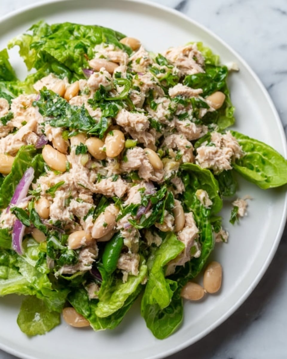 The image shows a fresh salad served on a white plate placed on a white marbled surface. The salad has three visible layers: the bottom layer is made of green leafy lettuce with a crisp texture, the middle layer contains chunks of light beige tuna mixed with finely chopped green herbs, and the top layer is scattered with light cream-colored beans and small pieces of purple onion, adding color and texture. The salad looks fresh and lightly mixed, with no dressing visible. Photo taken with an iphone --ar 4:5 --v 7