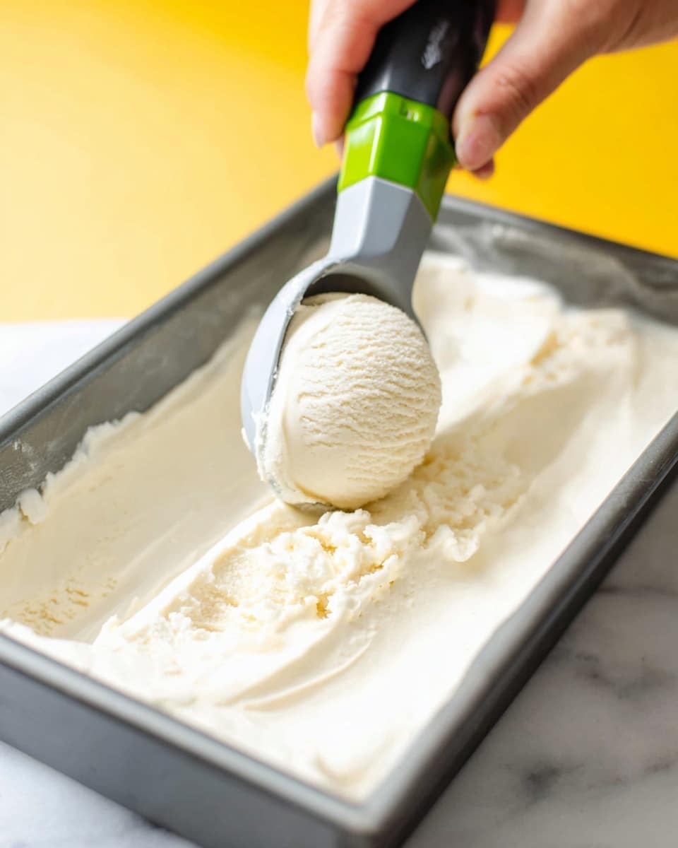 A close-up image shows a scoop of smooth, creamy off-white ice cream being lifted by a gray and green ice cream scooper inside a rectangular gray metal container filled with the same ice cream. A woman's hand holds the scooper on the left side of the photo, and the background is a blurred yellow color. The ice cream has a soft, thick texture and appears fresh and rich. The image is captured on a white marbled surface. photo taken with an iphone --ar 4:5 --v 7