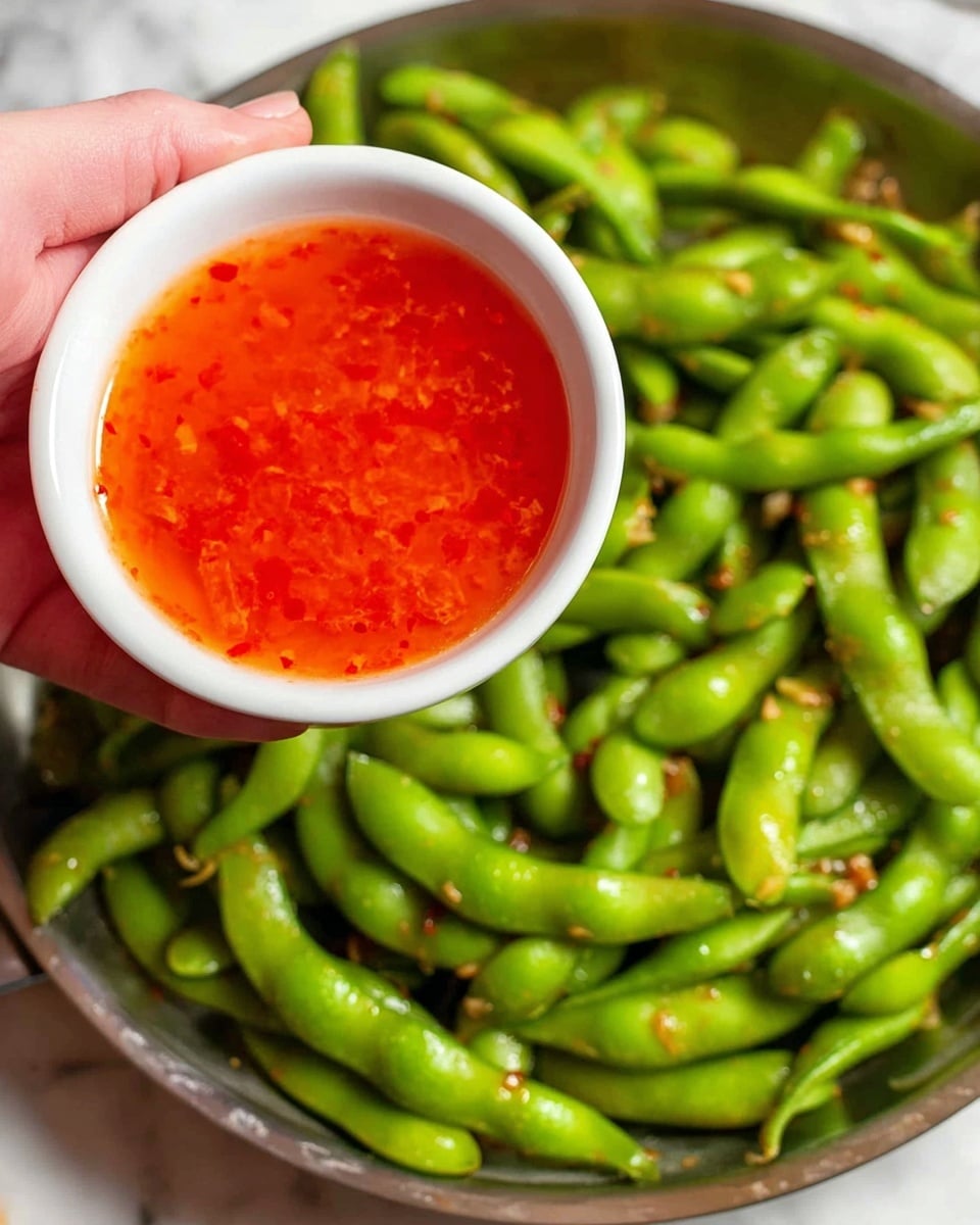 A woman's hand is holding a small white bowl filled with bright red-orange sauce that has small red bits in it. Below the bowl, there is a large silver pan full of shiny green edamame pods mixed with small bits of garlic or seasoning. The background shows a white marbled surface. The image is close up, showing the details of the sauce and the texture of the edamame pods photo taken with an iphone --ar 4:5 --v 7