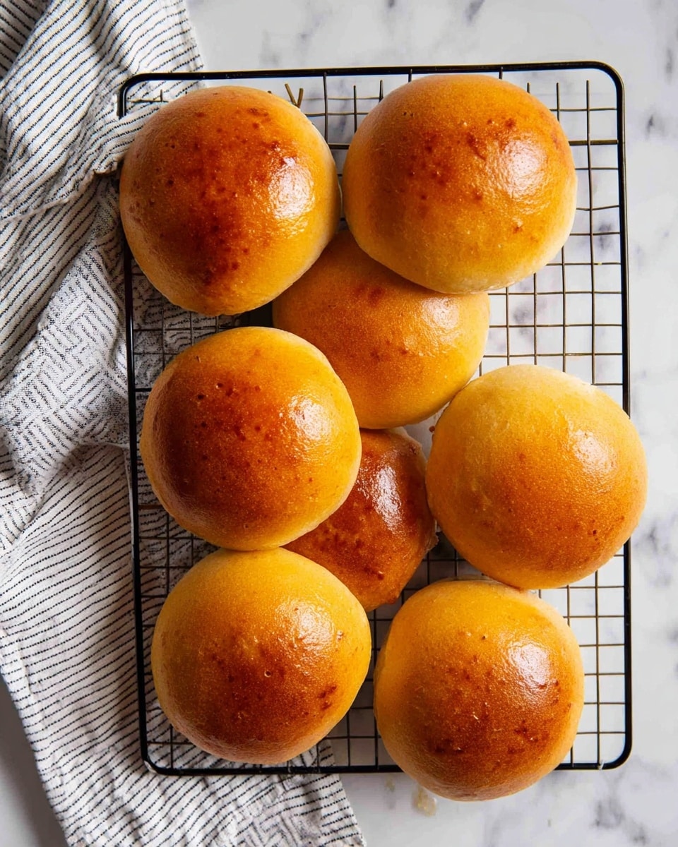Eight round golden brown buns with shiny smooth tops are arranged on a black cooling rack. The buns have a soft, slightly puffy texture with even coloring. To the right side, a white and gray striped cloth is partly visible on a white marbled surface under the rack. photo taken with an iphone --ar 4:5 --v 7