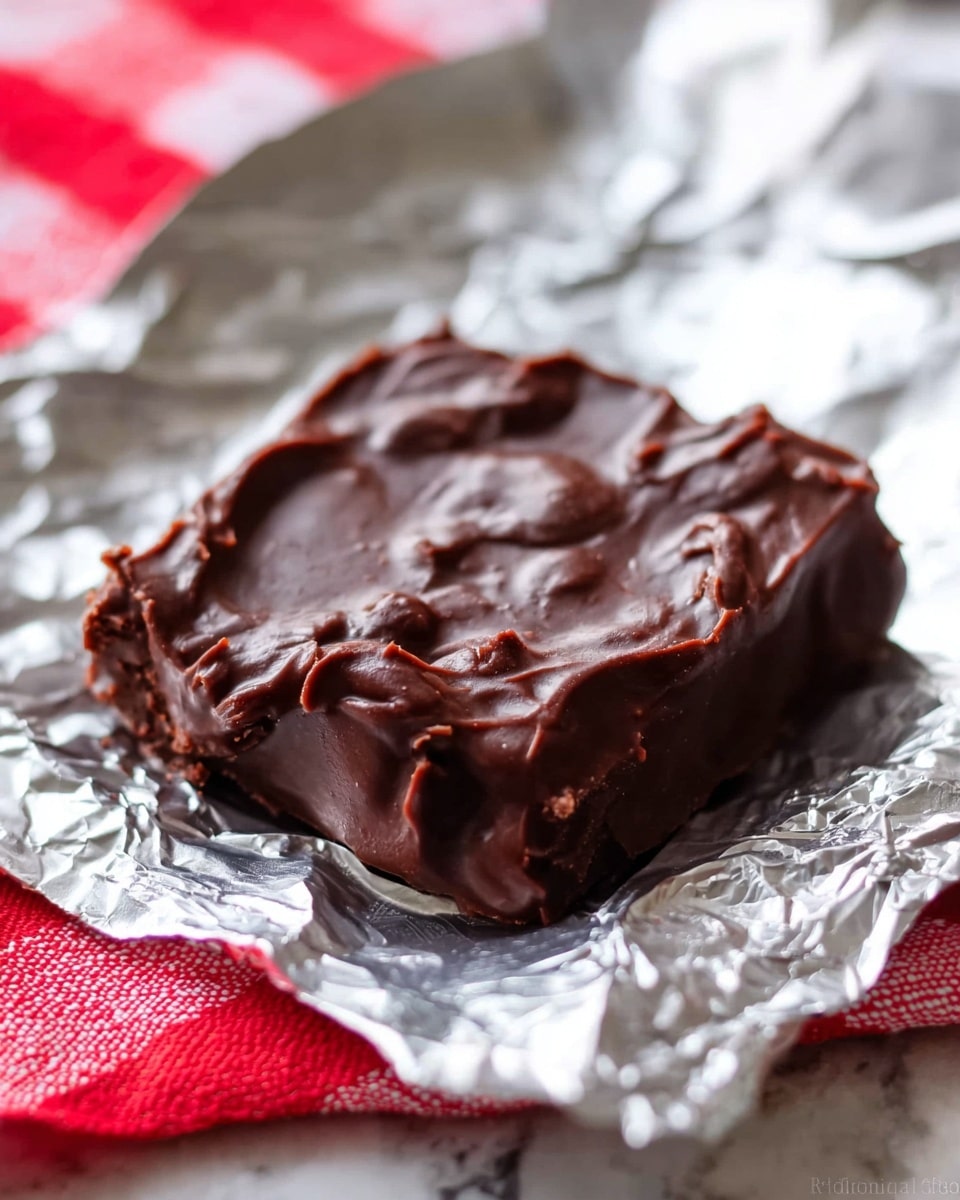 A single uneven square piece of dark chocolate fudge sits on shiny crinkled silver foil, showing a glossy, creamy texture with small lumps and swirls on top. The background has a soft blurry touch of red and white checkered fabric at the lower left corner, all placed on a white marbled surface. Photo taken with an iphone --ar 4:5 --v 7