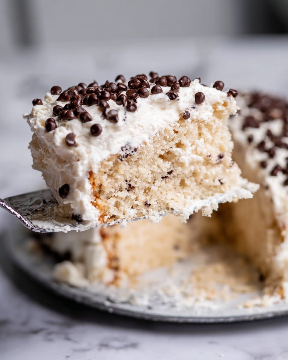 A close-up of a slice of light tan cake with a crumbly texture, topped with a thick layer of white creamy frosting sprinkled with small, dark brown chocolate chips, all held up by a silver server above a white marbled surface with more of the frosted cake visible in the background, photo taken with an iphone --ar 4:5 --v 7