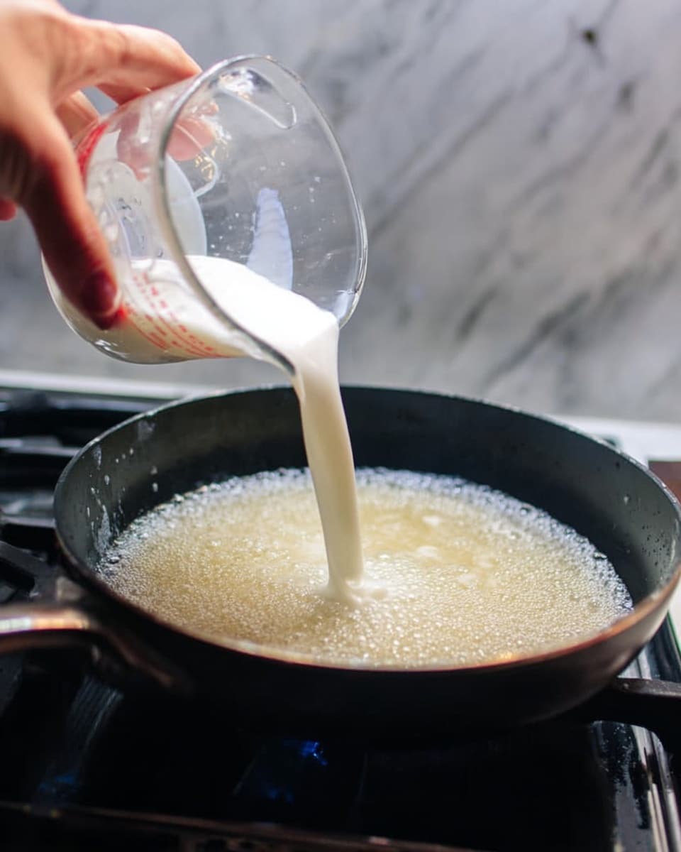 A woman's hand is pouring white milk from a clear glass measuring cup into a black pan filled with bubbling light golden liquid, sitting on a stove burner. The background features a white marbled surface. Photo taken with an iphone --ar 4:5 --v 7