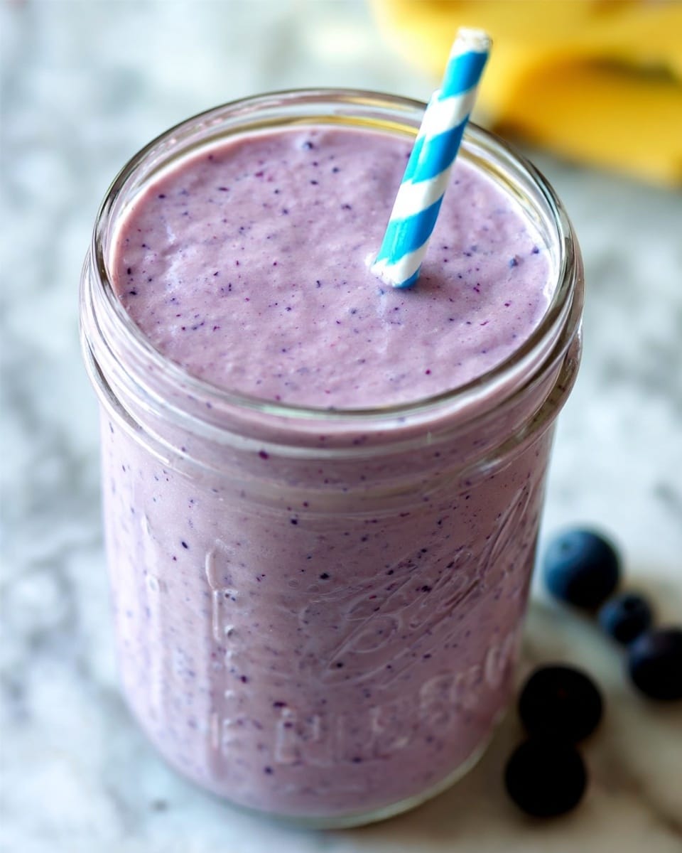 A metal measuring cup filled with many dark purple blueberries, showing their round shape and smooth texture. The cup is held by a woman's hand on the left side of the image. The background is softly blurred with warm light tones, placed on a white marbled surface. The blueberries fill the cup to the top and appear fresh and plump. photo taken with an iphone --ar 4:5 --v 7