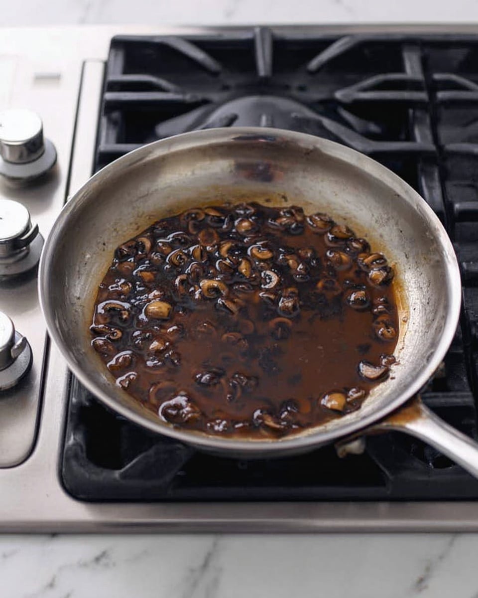 The image shows a metal frying pan on a stove burner. Inside the pan is a dark brown sauce with small chunks of mushrooms evenly spread throughout the liquid. The pan's surface has some slight discoloration from cooking. The stove is a shiny metallic color with black grates and silver knobs visible on the left side. The background surface beneath the stove is a smooth white marbled texture. photo taken with an iphone --ar 4:5 --v 7