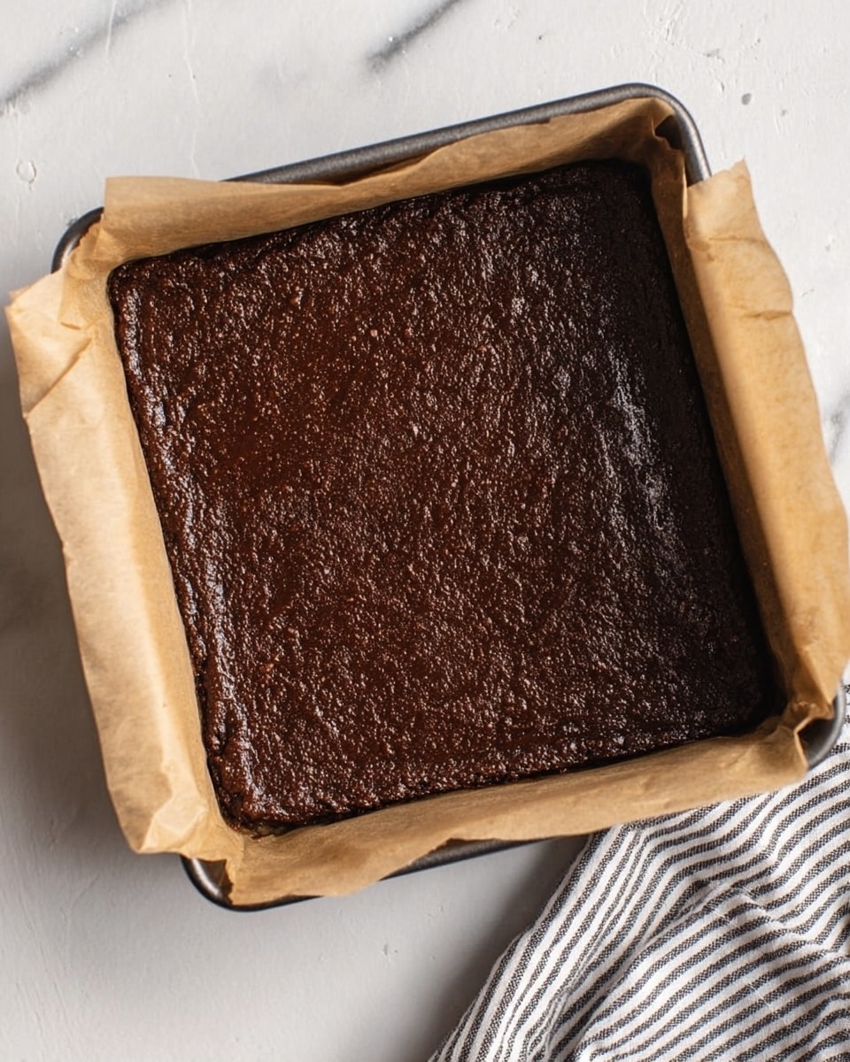 A square baking pan lined with brown parchment paper contains a thick, dark brown chocolate dessert with a smooth but slightly textured surface that fills the entire pan evenly. The pan is placed on a white marbled surface, and a striped cloth is partially visible in the bottom right corner. Photo taken with an iphone --ar 4:5 --v 7