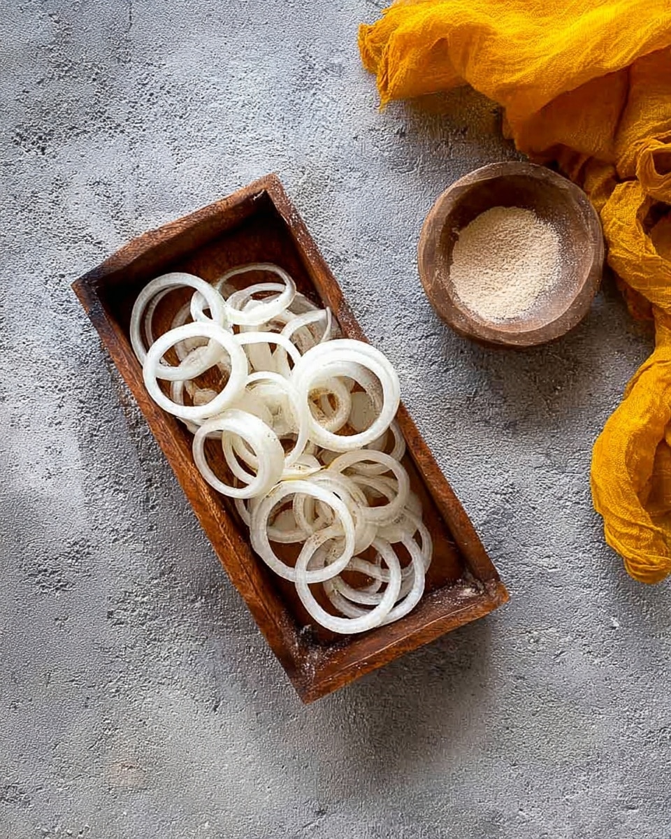 The image shows a rectangular wooden tray filled with several white onion rings scattered in a loose pile, some overlapping each other, on a gray rough textured surface. Above the tray, a small wooden bowl containing a light brown powdery substance is partially visible on the right side, and a bright yellow fabric is placed near the top left corner. The overall scene has a simple rustic feel with a mix of natural earthy tones and a slight dusting of powder on the tray among the onion rings photo taken with an iphone --ar 4:5 --v 7