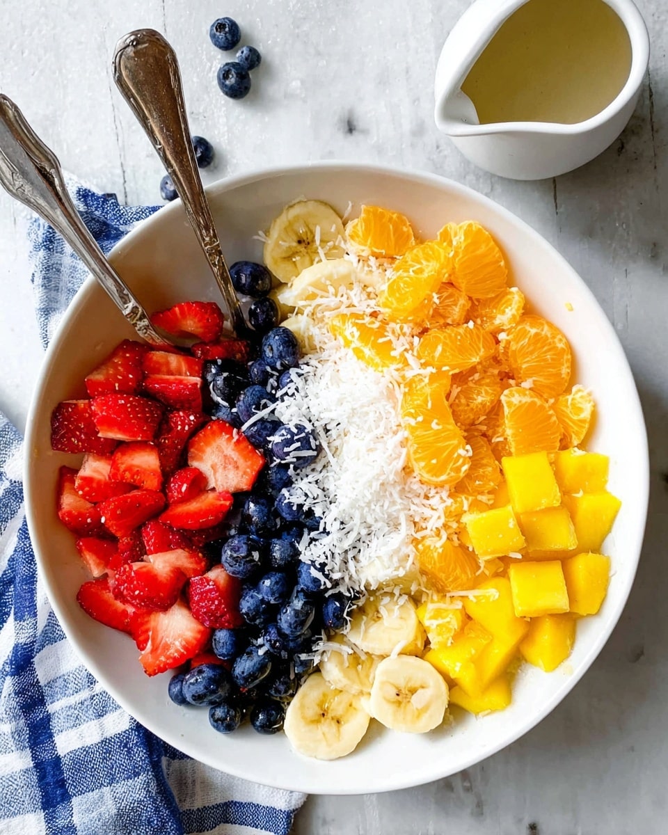 A white bowl filled with six colorful layers of fruit arranged side by side: red sliced strawberries on the top left, bright orange mandarin orange segments on the top right, shredded white coconut in the middle, deep blue fresh blueberries scattered over several layers, pale yellow sliced bananas on the lower right, and bright yellow diced mango pieces on the lower left. Two silver spoons rest inside the bowl, one near the strawberries and the other near the mango. The bowl sits on a white marbled surface with a small white sauce pitcher nearby and a blue checked cloth partially visible. Photo taken with an iphone --ar 4:5 --v 7