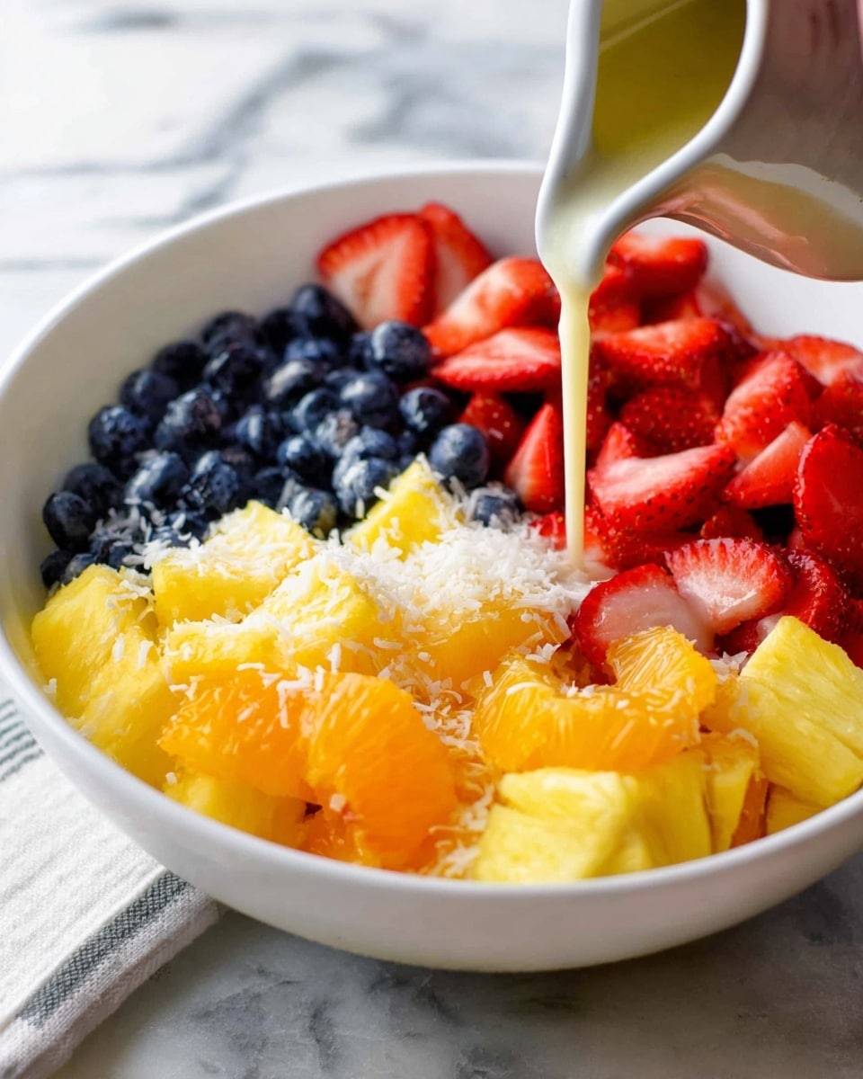 A white bowl holds a vibrant fruit salad with four visible layers: bright red sliced strawberries with a smooth texture in the top left, deep blue blueberries with a smooth but slightly glossy surface in the top right, juicy yellow pineapple chunks with a fibrous texture on the left bottom half, and soft orange mandarin pieces with a slightly shiny surface on the bottom right. There is a sprinkling of white shredded coconut scattered on top of all the fruits. A white ceramic pitcher pours a light cream-colored sauce over the pineapple in the center. The bowl sits on a white marbled surface, and a woman's hand is partially visible holding the pitcher. Photo taken with an iphone --ar 4:5 --v 7