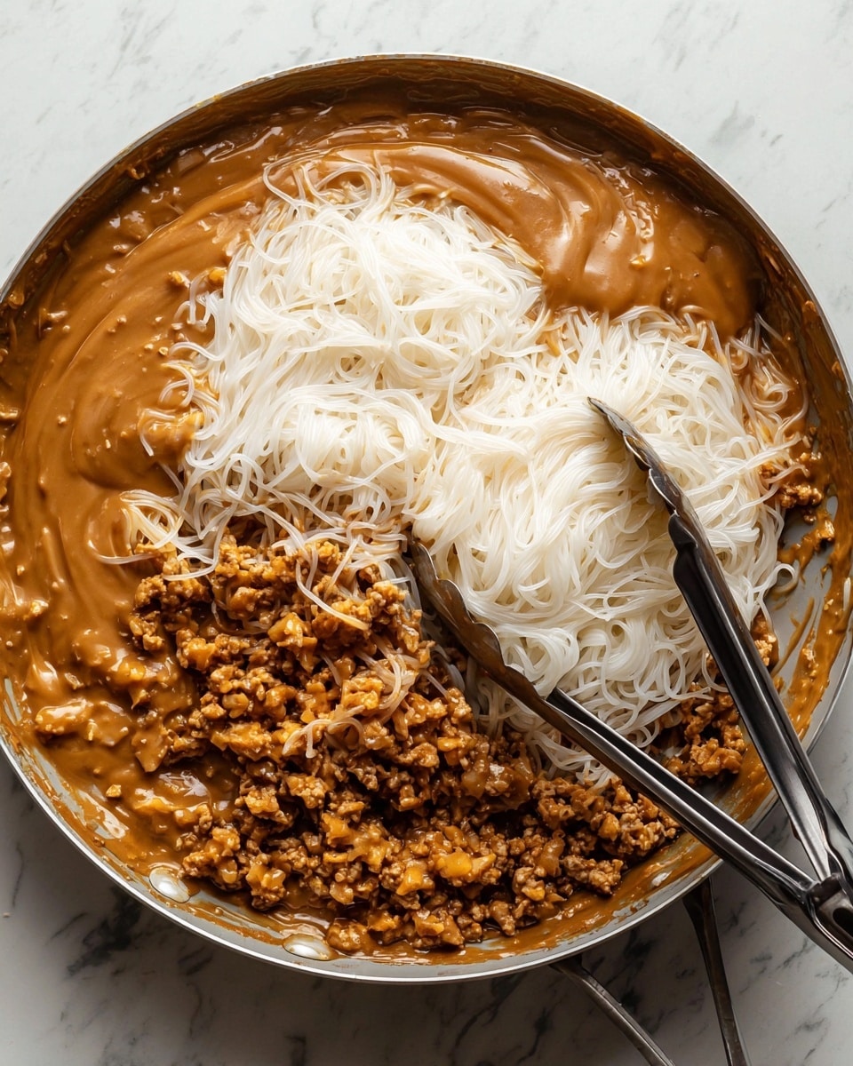 The image shows a pan with three main layers: at the bottom, there is a layer of cooked, crumbled light brown meat with some darker browned spots. Above this, there is a large portion of thin, white rice noodles sitting loosely in strands. The top layer is a thick, smooth brown sauce covering part of the noodles and meat, blending slightly at the edges. A pair of metal tongs with black handles rests on the noodles and meat at the bottom right corner of the pan. The pan sits on a white marbled surface. Photo taken with an iphone --ar 4:5 --v 7