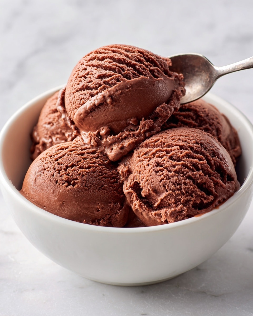 A white bowl filled with four scoops of rich, creamy chocolate ice cream layered closely together, each scoop showing a smooth, slightly textured surface with a deep brown color. A spoon is lifting a scoop from the side, capturing the soft, thick texture of the ice cream with small ridges and folds. The bowl sits on a white marbled surface, giving a clean and simple background. photo taken with an iphone --ar 4:5 --v 7