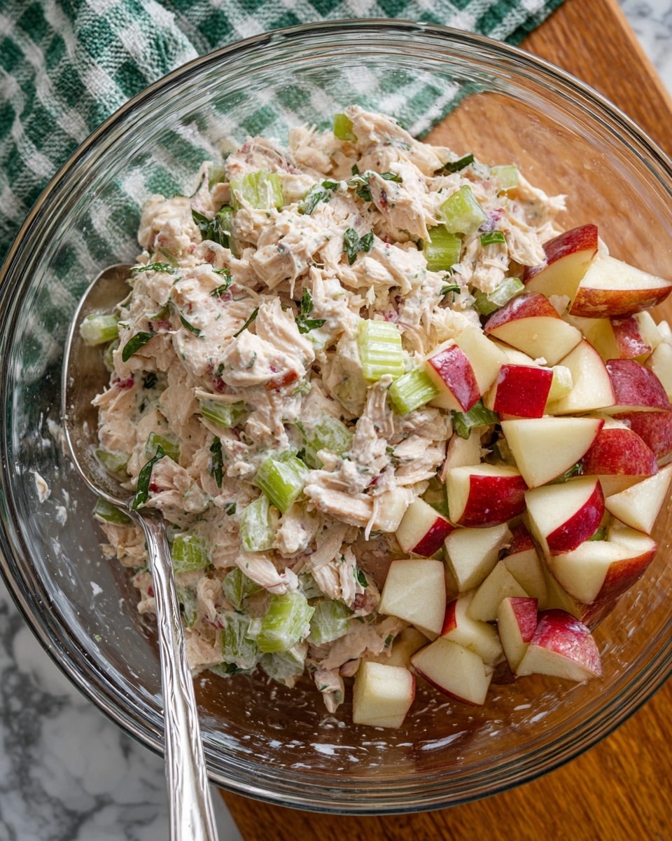A clear glass bowl filled with a mix of light pink shredded chicken combined with small light green celery pieces, fresh herbs, and diced pieces of apple with red skin, which are piled on one side of the bowl. The chicken mixture has a creamy texture, and the apple pieces have a firm, crisp look with red and white colors. A silver spoon rests inside the bowl near the bottom left. The bowl sits on a wooden surface with a green and white checkered cloth in the background, but the overall setting should be imagined on a white marbled surface. photo taken with an iphone --ar 4:5 --v 7