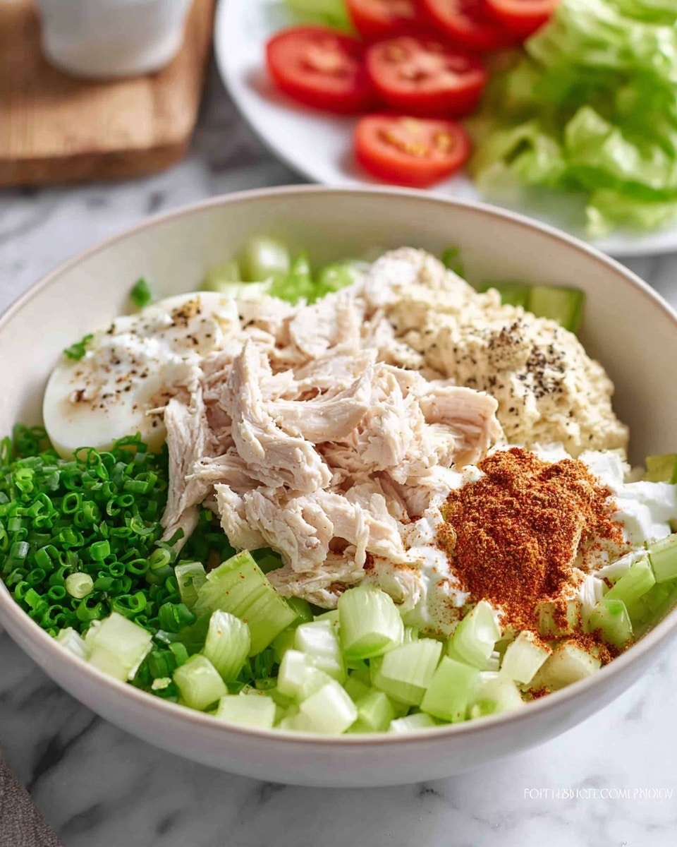 The image shows a white bowl filled with several layered ingredients arranged separately inside. The bottom layers are light green chopped celery pieces and finely chopped green chives scattered throughout. On top of the celery next to the chives is shredded white cooked chicken seasoned with black pepper. Next to the chicken is a creamy white mayonnaise layer with a small pile of reddish-brown spice powder and a dollop of coarse grainy mustard. In the background, there is a partially visible white plate with green lettuce and sliced red tomatoes on top. The whole scene is set on a white marbled surface. Photo taken with an iphone --ar 4:5 --v 7