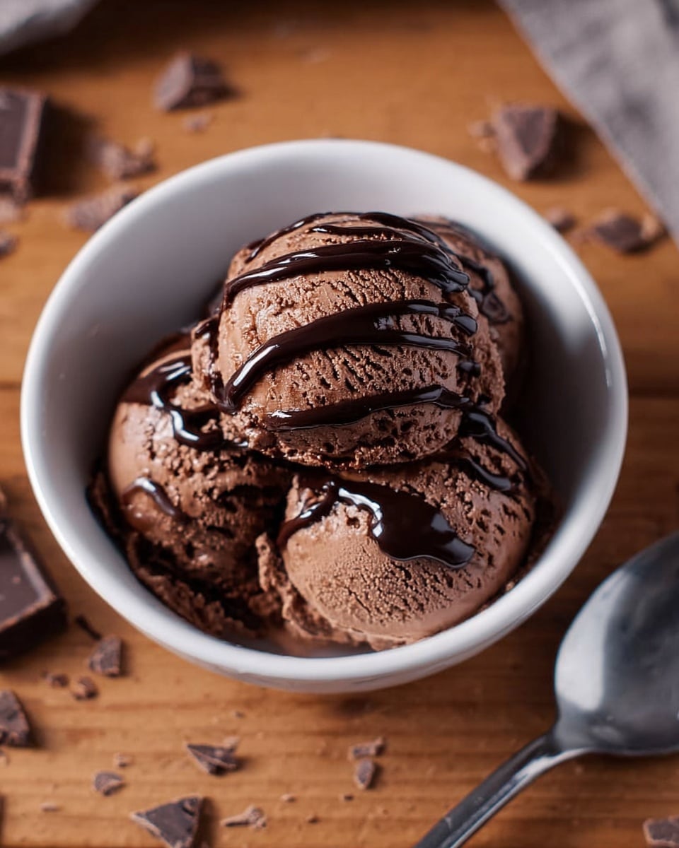 A white bowl contains three round scoops of chocolate ice cream, each scoop showing a creamy, slightly rough texture with visible air pockets. The top scoop is drizzled with dark, shiny chocolate syrup that runs down in thin, uneven lines. The bowl sits on a wooden surface with scattered chocolate pieces around it, and a metal spoon is placed to the right side. photo taken with an iphone --ar 4:5 --v 7