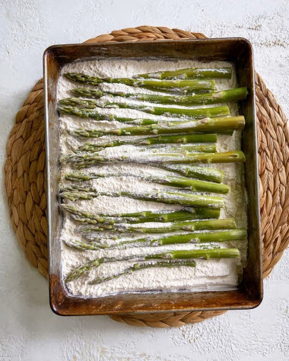 A rectangular metal tray is filled with a layer of white flour spread unevenly across the bottom and sides. On top, a single layer of green asparagus spears is placed horizontally, each partially covered with white flour, creating a mix of green and white textures. The tray sits on a round woven mat, and the background is a white marbled surface. photo taken with an iphone --ar 4:5 --v 7