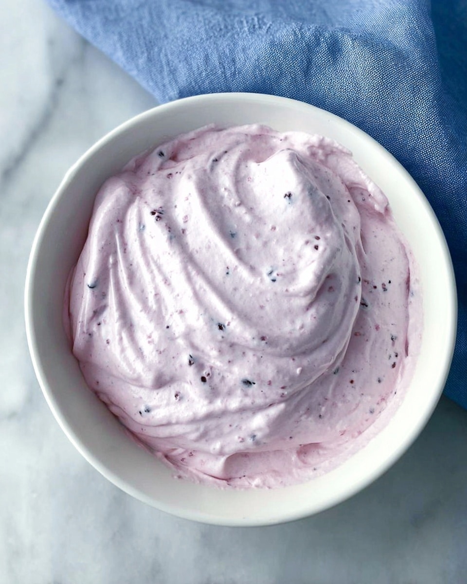 A white bowl filled with a single thick layer of light purple whipped mixture that is smooth with some small darker purple bits scattered throughout, sitting on a white marbled textured surface with a corner of a blue cloth visible behind the bowl, photo taken with an iphone --ar 4:5 --v 7