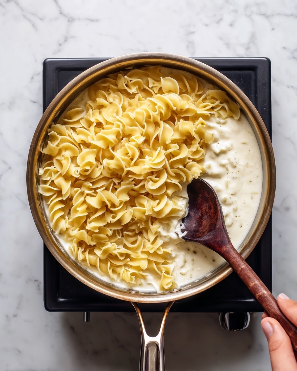 A close-up top view of a silver pan on a small black stove with creamy white sauce at the bottom layer, and twisted pale yellow pasta covering nearly all the sauce on top, with a dark wooden spoon stirring the pasta from the right side, held by a woman's hand. The pan is on a white marbled surface. Photo taken with an iphone --ar 4:5 --v 7