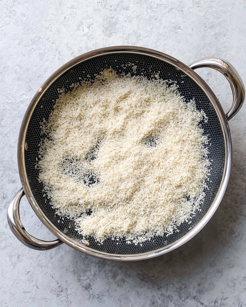 A round, deep pan with silver handles on a white marbled surface holds a thin layer of uncooked white rice grains spread unevenly across the bottom. The pan’s interior is dark with a subtle honeycomb pattern, contrasting with the pale rice scattered inside. The silver handles curve outward, adding a sleek metallic touch to the scene. Photo taken with an iphone --ar 4:5 --v 7