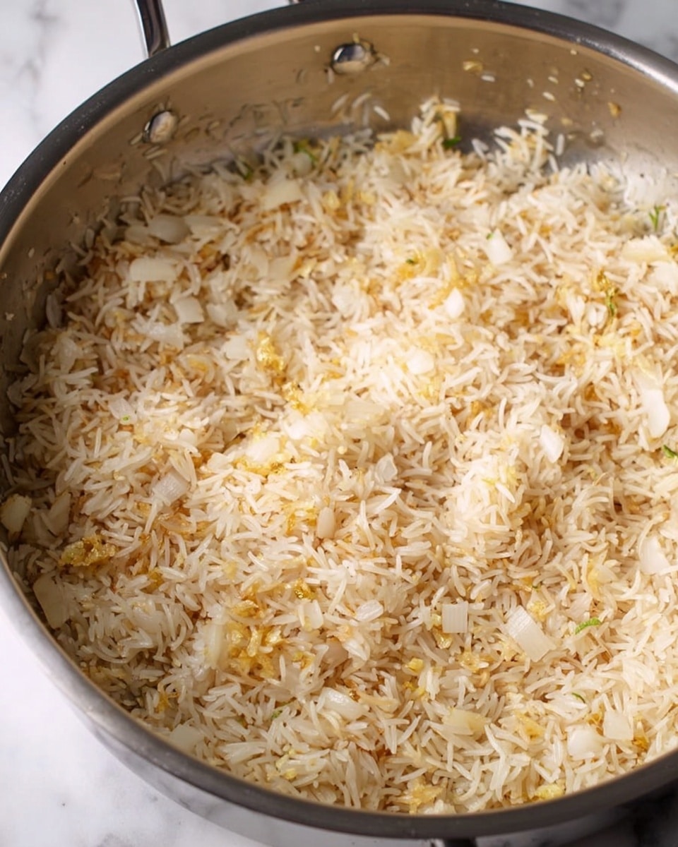 A close-up view shows a silver metal pan filled with a mixture of small, thin rice grains and finely chopped onions. The rice is light beige in color and evenly spread across the pan, mixed with small pieces of white onion and some yellow bits, likely garlic or ginger, scattered throughout. The texture looks slightly sautéed but still dry, with the ingredients blending together in a rough, uneven layer. The pan’s shiny inner surface contrasts softly with the pale food inside. The background and surface are a white marbled texture. photo taken with an iphone --ar 4:5 --v 7