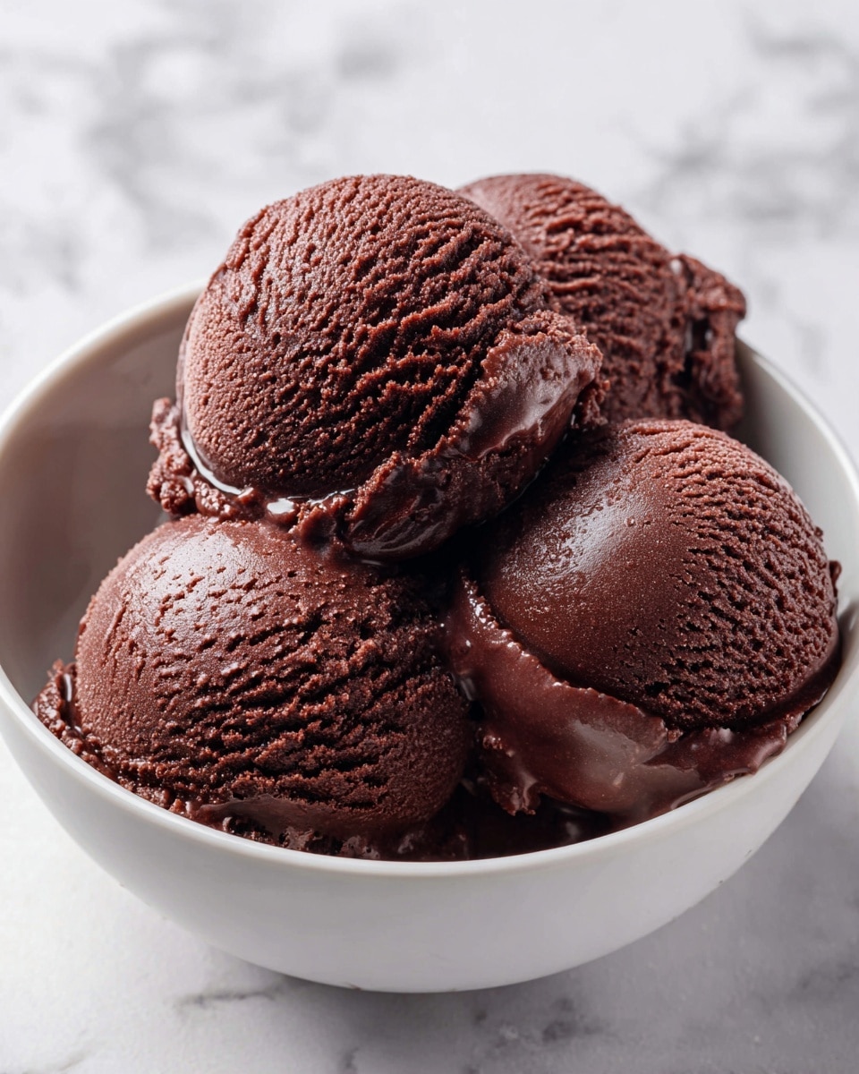 The image shows four scoops of chocolate ice cream placed close together on a white bowl with a smooth inside. The ice cream is dark brown with a textured surface, having visible small ridges and soft bumps that make it look creamy and cold. There is a slight melting effect around the edges of the scoops, giving a shiny wet look in spots. The background is a white marbled texture which contrasts with the deep brown ice cream. photo taken with an iphone --ar 4:5 --v 7