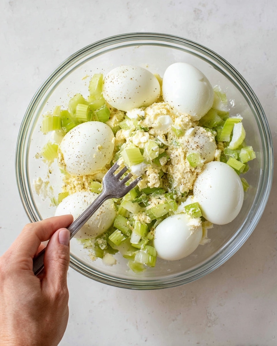 A clear glass bowl sits on a white marbled surface, containing four whole white hard-boiled eggs along with chopped pale green celery scattered around them. There is a creamy, white base with specks of black pepper visible under and around the eggs and celery. A woman's hand is holding a silver fork, pressing down and mashing a few of the eggs, with some pieces broken and mixed with the celery and creamy mixture. photo taken with an iphone --ar 4:5 --v 7