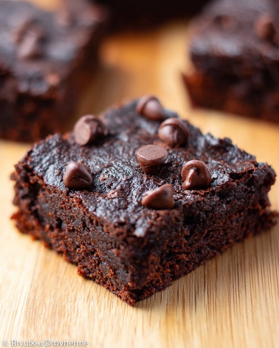 A close-up view of a single square brownie with a rough, rich dark brown texture that looks moist and dense. The top layer is decorated with several shiny dark chocolate chips, some slightly melted and others firm, scattered unevenly. The brownie sits on a light wooden surface with a soft focus on another similar brownie piece in the background. The overall look highlights the thick, chunky chocolate treat with a slight crumbly edge. Photo taken with an iphone --ar 4:5 --v 7